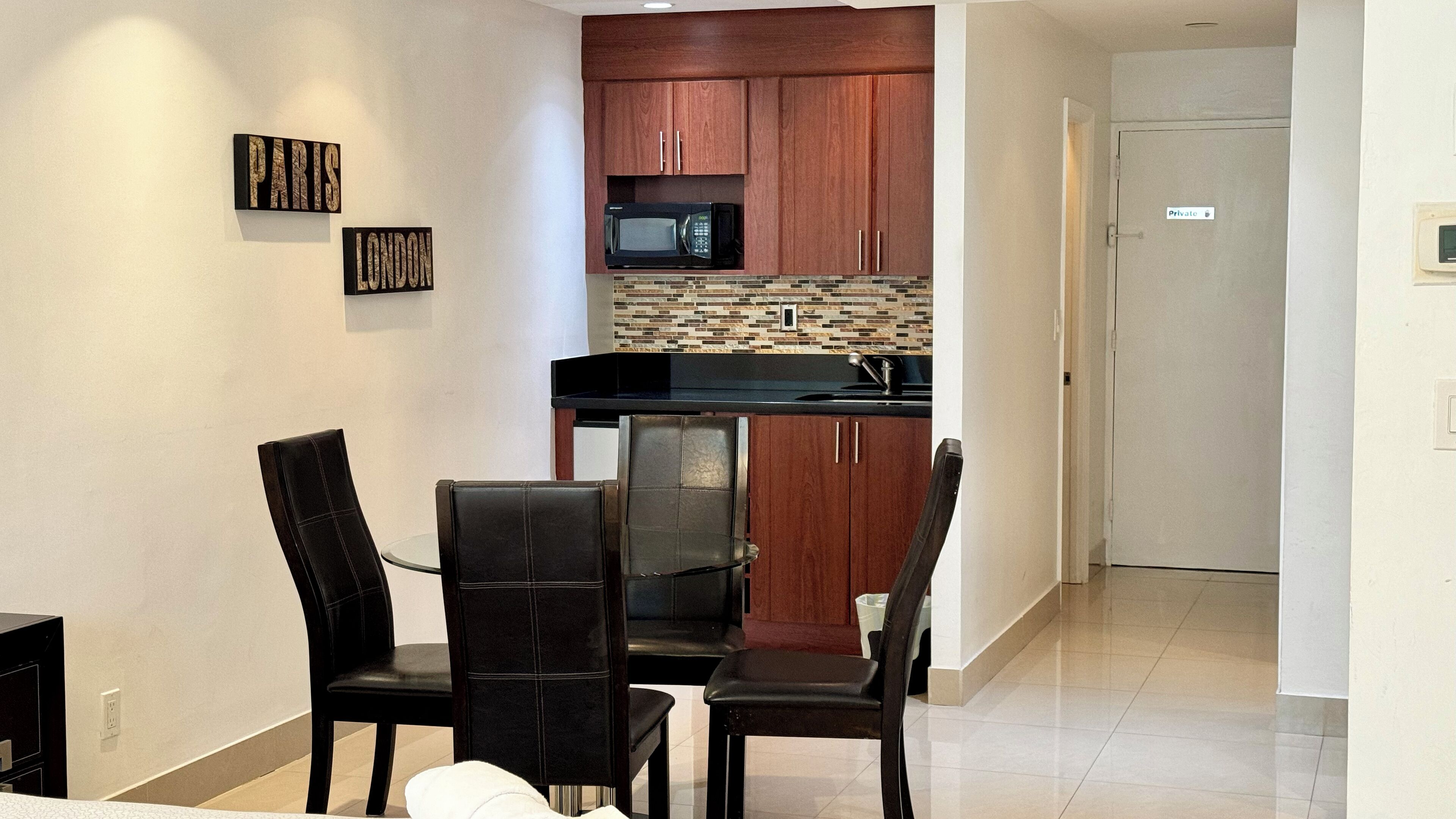 A compact kitchen with wooden cabinets, a microwave, and a tiled backsplash. There is a dining area with two chairs and a table.