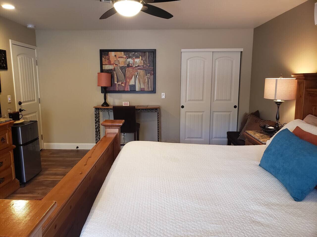 A bedroom with a bed, a desk, a chair, a lamp, and a painting on the wall.