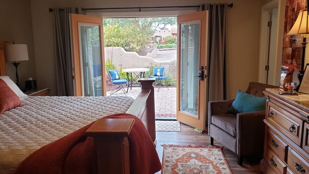 A hotel room with a bed, a dresser, a chair, and a view of an outdoor patio with chairs and a table. - Robin`s Red Rock Getaway Suite A (Sedona)