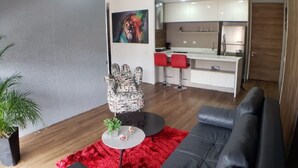 Una sala de estar moderna con un sofá de cuero negro, un tapete rojo, un sillón estampado y una pequeña mesa redonda con una planta y dos vasos.