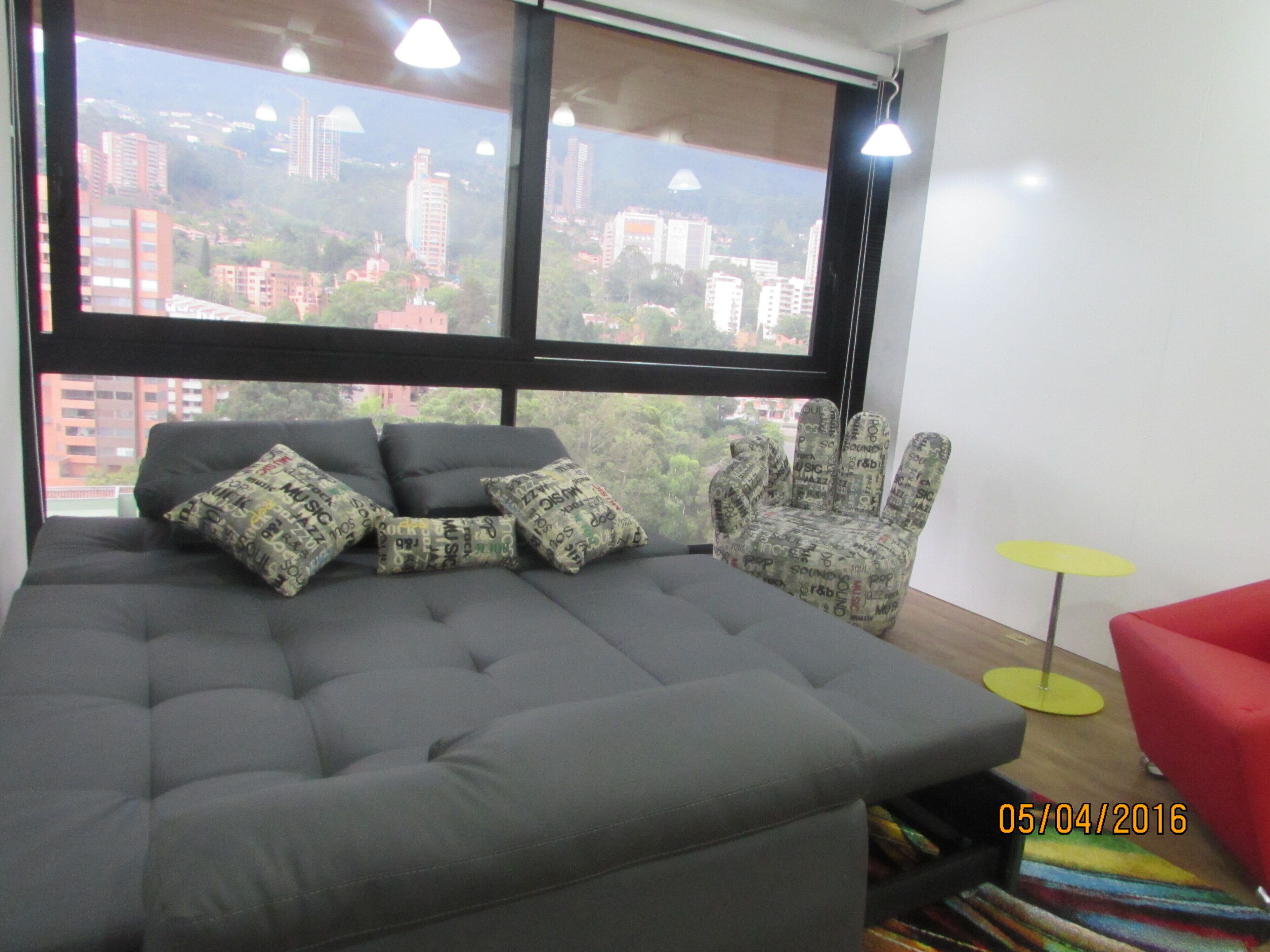 A modern living room with a grey sofa, patterned armchair, and a red chair. A city view is visible through large windows.