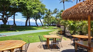 A tropical outdoor dining area with wooden tables and chairs, a thatched-roof structure, and a view of the ocean. - Dine in paradise on your garden-view lanai (Lahaina)
