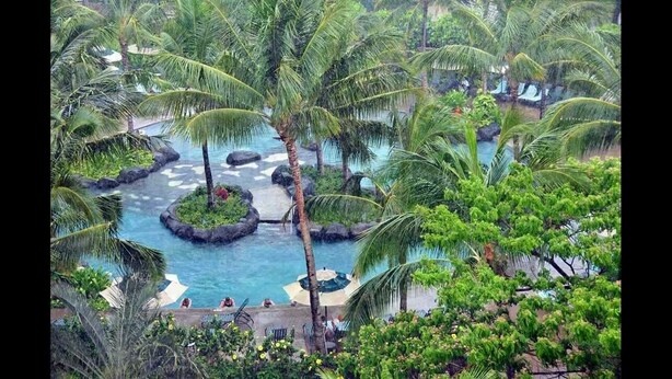 Un resort tropical con una piscina central rodeada de palmeras y formaciones rocosas.