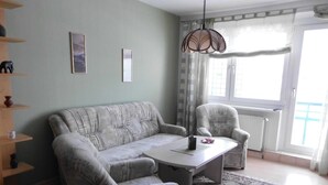A living room with a sofa, armchair, and a table. There is a patterned rug on the floor, a ceiling light fixture, and a window with curtains. - Apartment Bulang (Karlshagen)