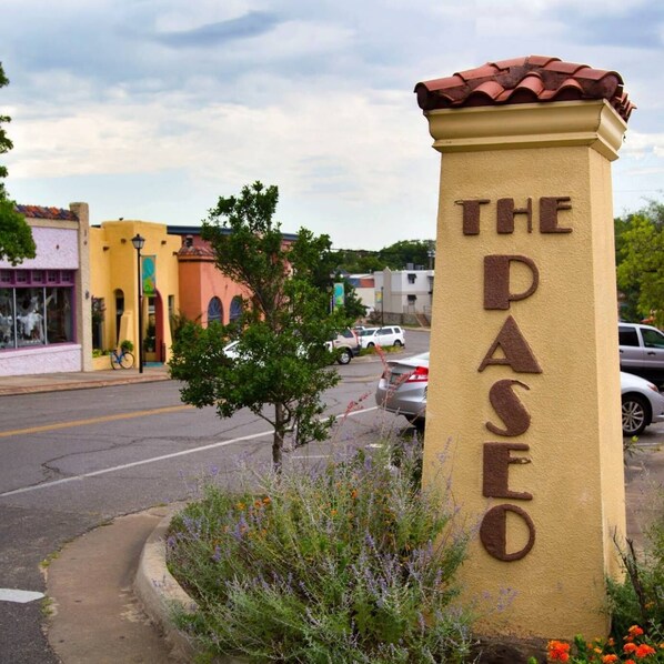 A sign with "THE PASEO" written on a beige pillar with a red roof, surrounded by greenery and a street view.
