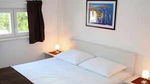 A neatly made bed with white linens, two bedside tables with lamps, a framed picture on the wall, and a window with curtains. - House Ercegovic A2 + 2 (Primošten)