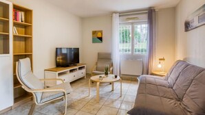 Uma sala de estar com sofá, duas poltronas, televisão, estante de livros, mesa com planta e janela com cortinas.