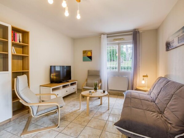 Una sala de estar con un sofá, dos sillones, un televisor, una estantería, una mesa con una planta y una ventana con cortinas.