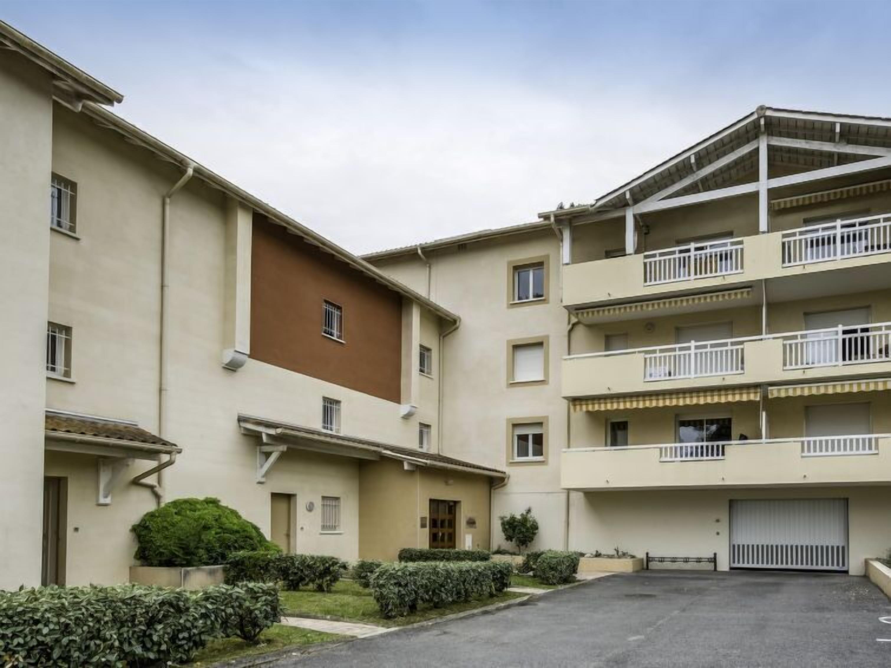 Um edifício residencial com vários andares, varandas, área de estacionamento e um pequeno jardim.
