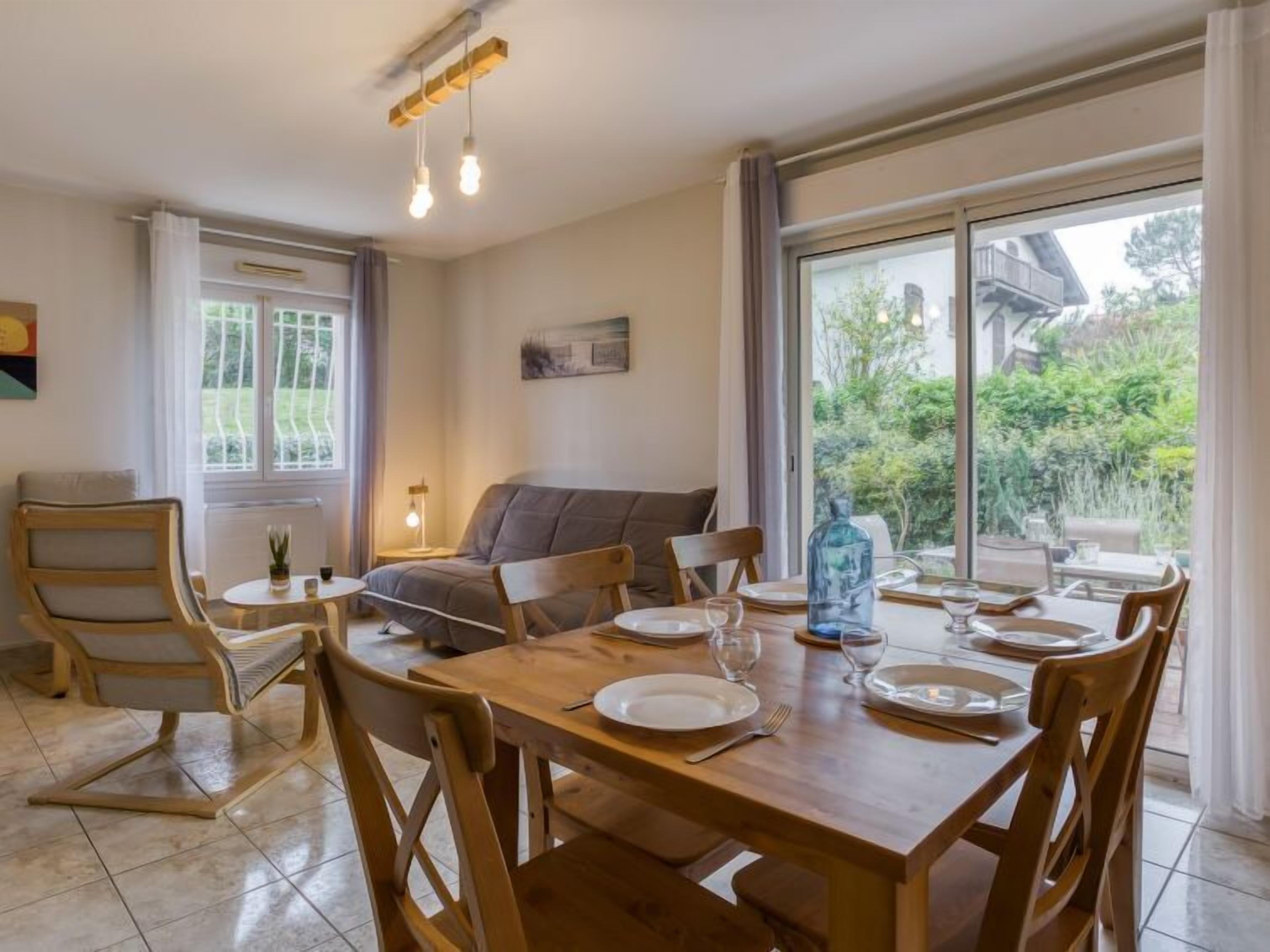 A dining area with a wooden table set for four, a grey sofa, and a window with a view of greenery.