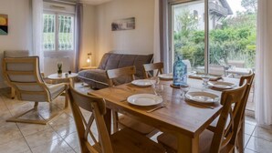 Uma sala de jantar com uma mesa de madeira posta para quatro pessoas, um sofá cinzento e uma janela com vista para o verde.