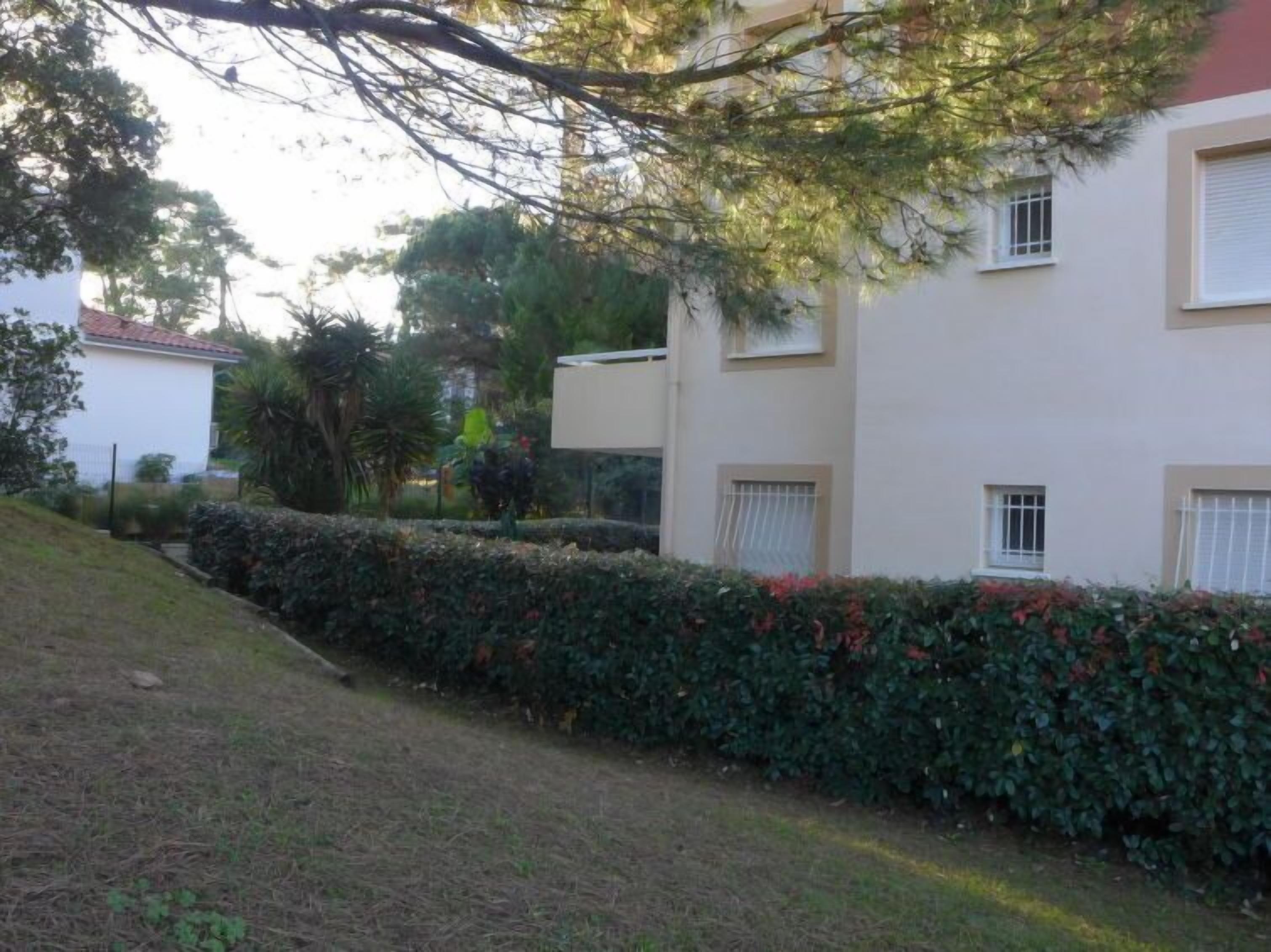 A multi-story building with a white exterior and a red roof. There are windows with white blinds, and a hedge with red flowers in front.
