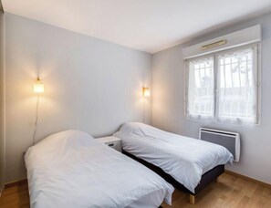 Una habitación sencilla y limpia con dos camas individuales, una mesita de noche pequeña, un ventanal con vistas y un calentador fijado a la pared.