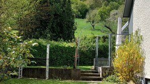 A well-maintained garden with a stone pathway leading to a gate, surrounded by lush greenery and a view of a hillside.