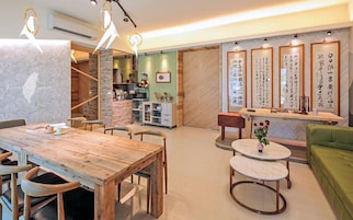 Un comedor moderno con una mesa y sillas de madera, una pequeña mesa redonda con un florero, y un sofá verde. Hay piezas de caligrafía enmarcadas en la pared y una zona de cocina al fondo.