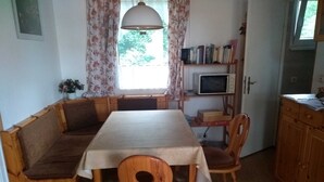 Ein Essbereich mit einer hölzernen Bank, einem Tisch und Stühlen. Ein Fenster mit Vorhängen, ein Bücherregal mit Büchern und eine Mikrowelle sind ebenfalls vorhanden.