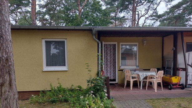 Ein kleines, eingeschossiges Gebäude mit gelber Fassade, einer überdachten Terrasse mit Tisch und Stühlen sowie einem kleinen Garten mit verschiedenen Pflanzen.
