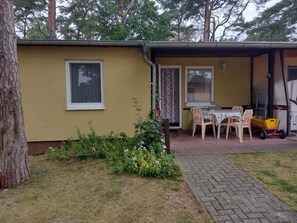 En liten, envåningsbyggnad med en gul fasad, en täckt uteplats med ett bord och stolar samt en liten trädgård med olika växter.
