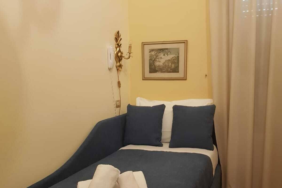 A single bed with a dark blue bedspread and white pillows, a framed picture on the wall, and a white curtain on the window.