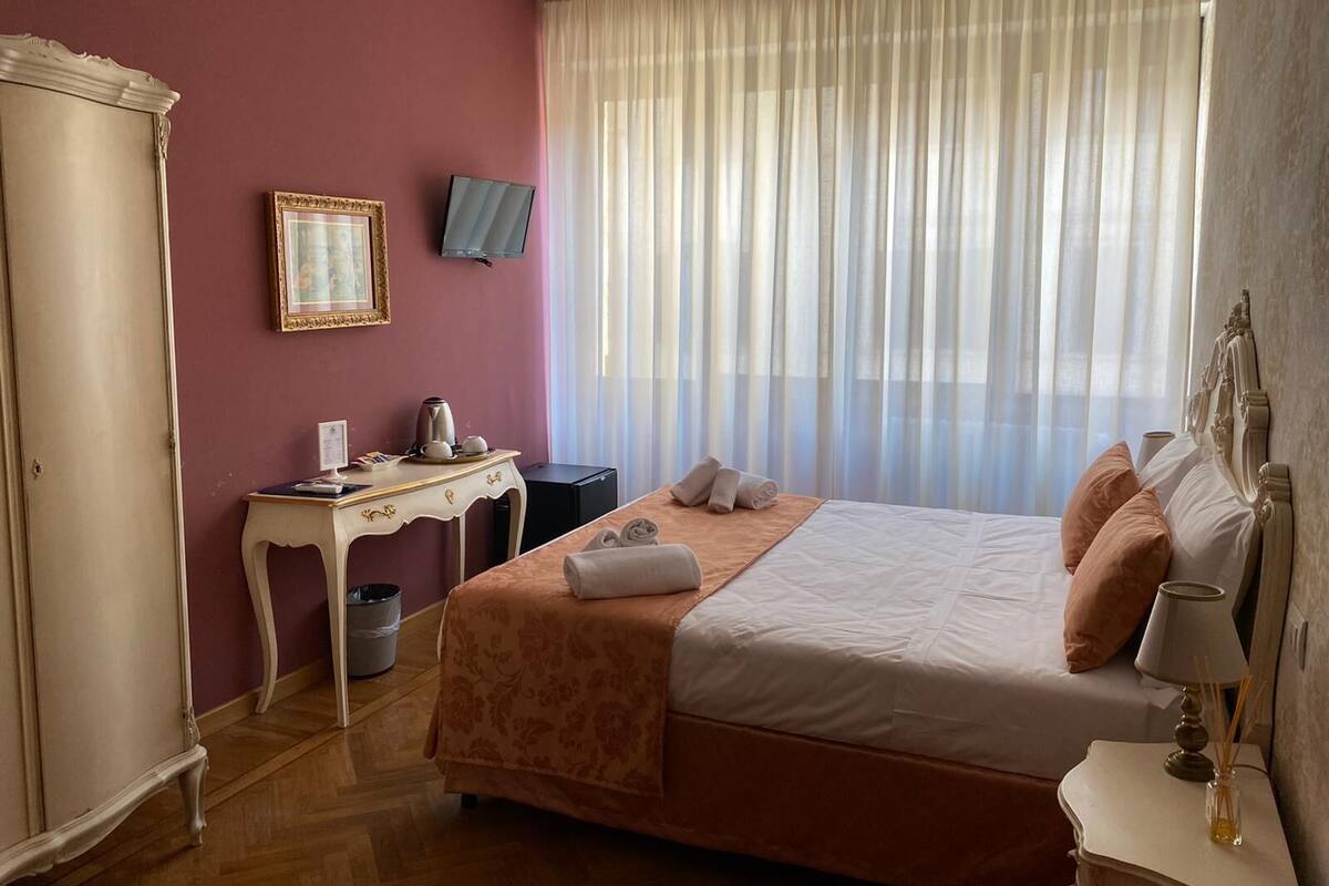 A hotel room with a bed, a desk, a television, and a large window with sheer curtains.