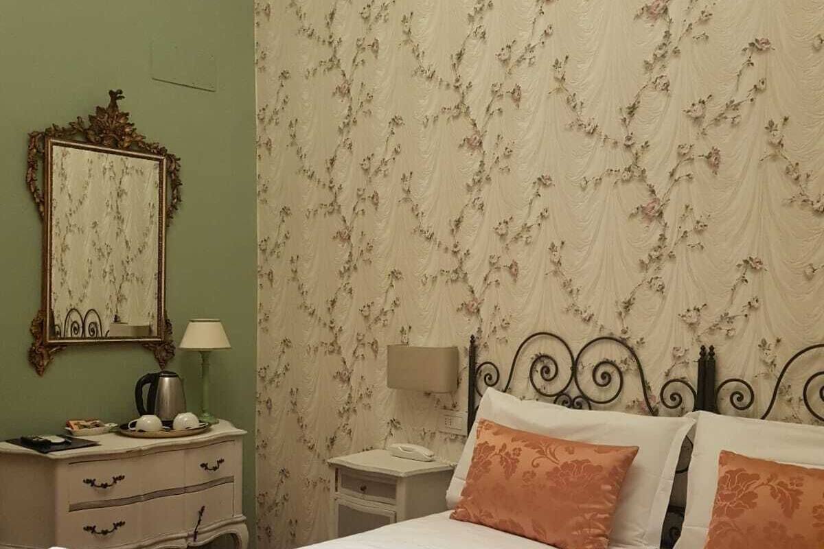 A hotel room with a bed, two bedside tables, a nightstand, a mirror, and a wall with floral wallpaper.