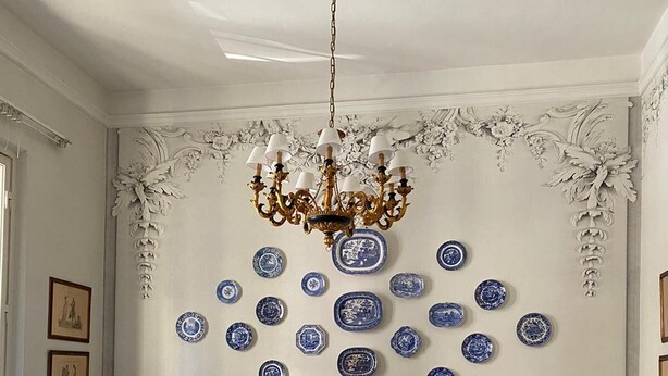 Un comedor con sillas y mesas blancas, una araña y platos decorativos azules y blancos en la pared.