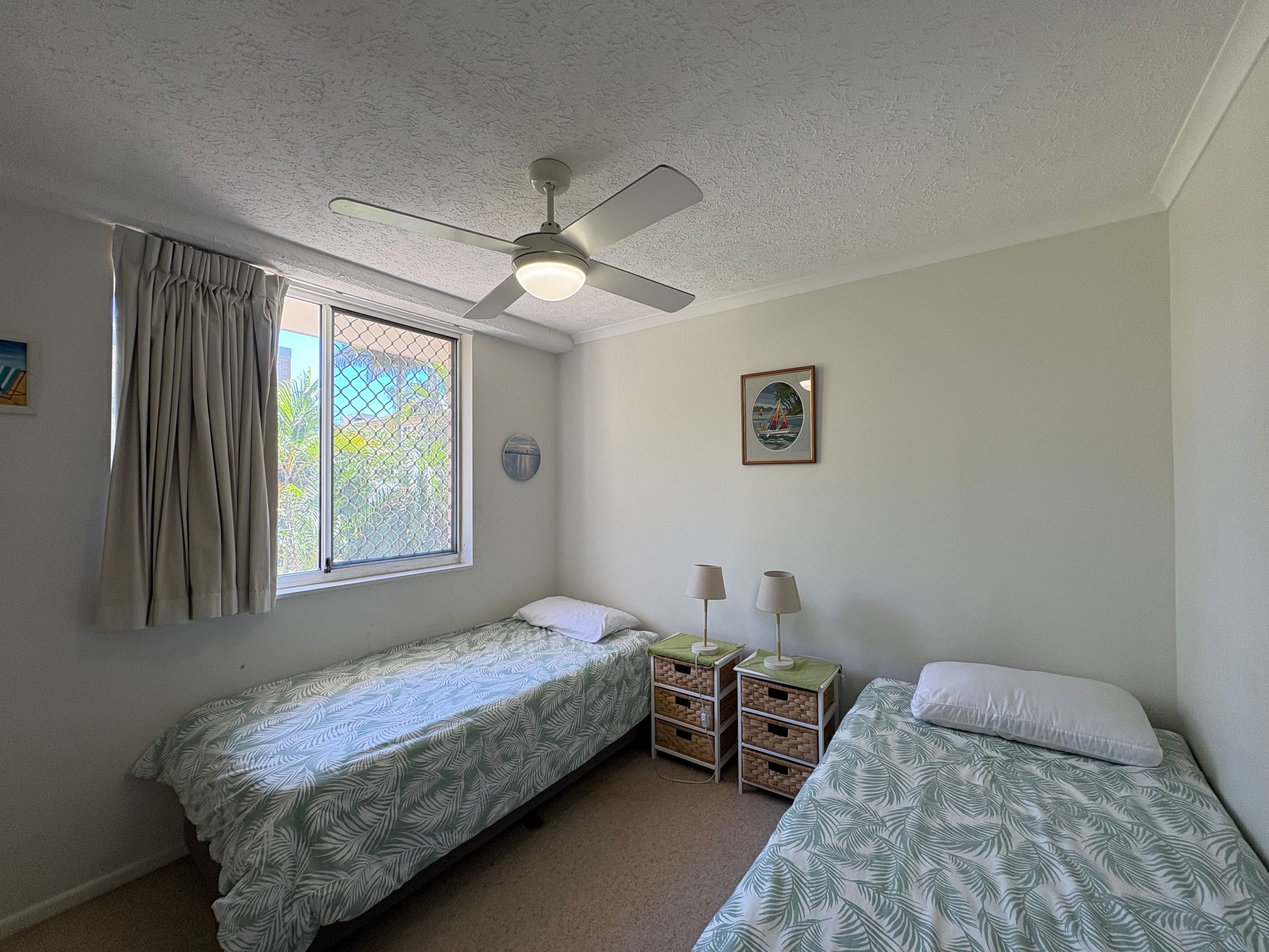 Une chambre avec deux lits simples, un ventilateur de plafond, une fenêtre avec des rideaux, une petite table avec des lampes et un tableau encadré au mur.