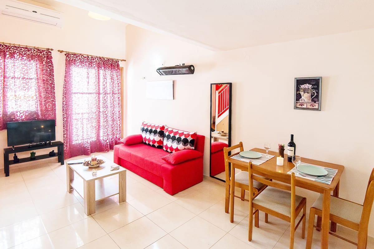 A living room with a red sofa, a coffee table, a TV, and a dining area with a table and chairs.