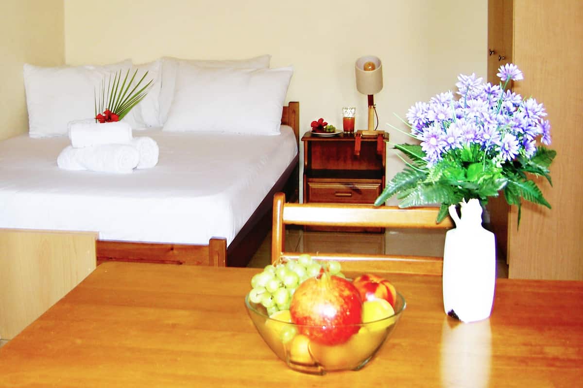 A hotel room with a neatly made bed, a bedside table with a lamp, a fruit bowl, and a vase with flowers.