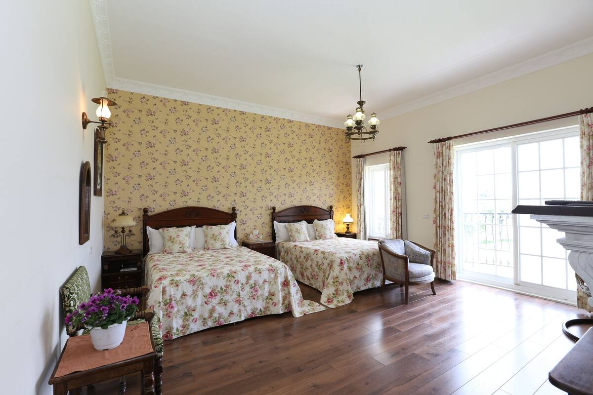 Un dormitorio con dos camas, una silla, una mesita de noche, una lámpara y una ventana con cortinas.