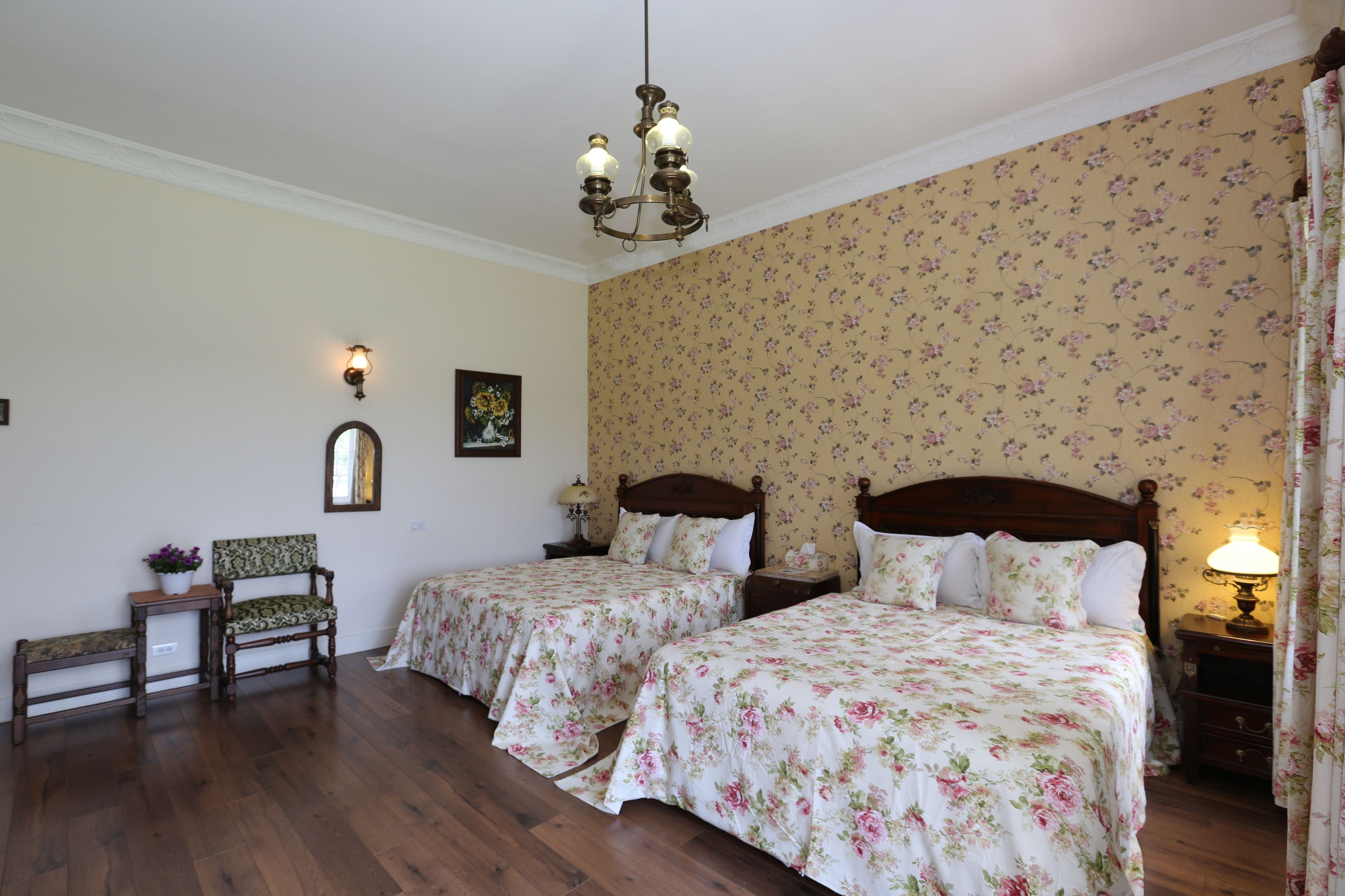 A bedroom with two beds, wooden flooring, floral wallpaper, a chandelier, a nightstand with a lamp, a chair, and a small table with a vase of flowers.