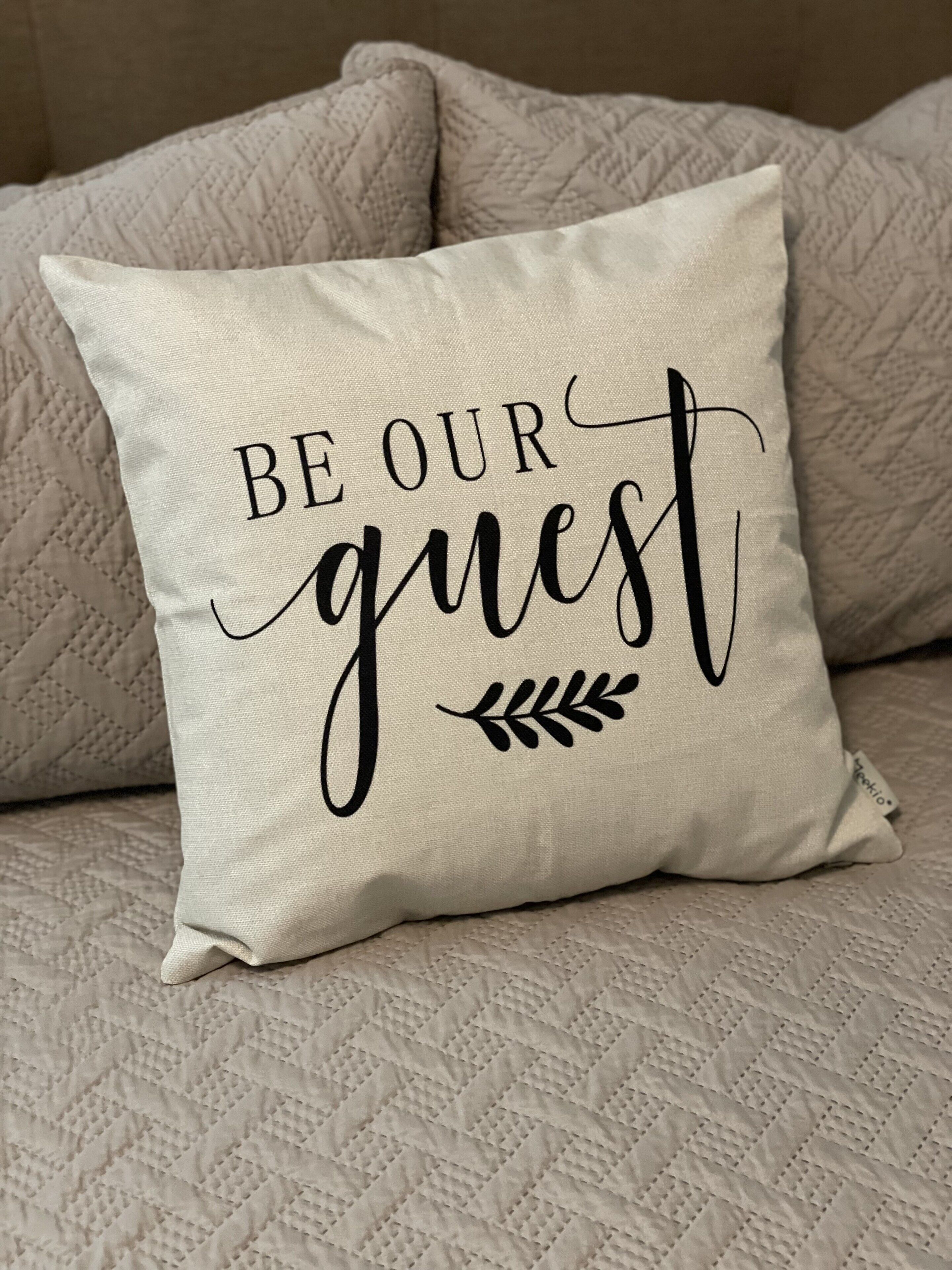 Een kussen met de tekst 'BE OUR guest' op een beige bank met een structuur.