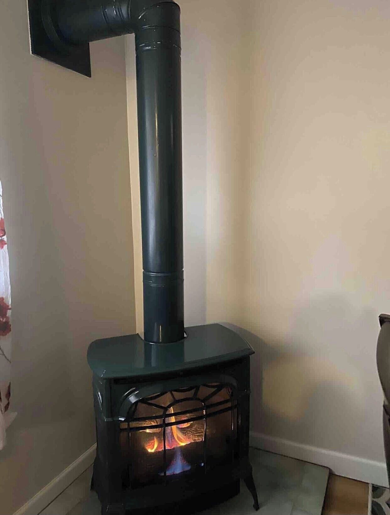 A wood-burning stove with a chimney, placed in a corner of a room with a plain wall and a tiled floor.