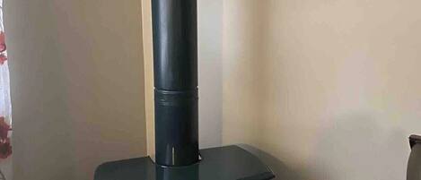 A wood-burning stove with a chimney, placed in a corner of a room with a plain wall and a tiled floor.