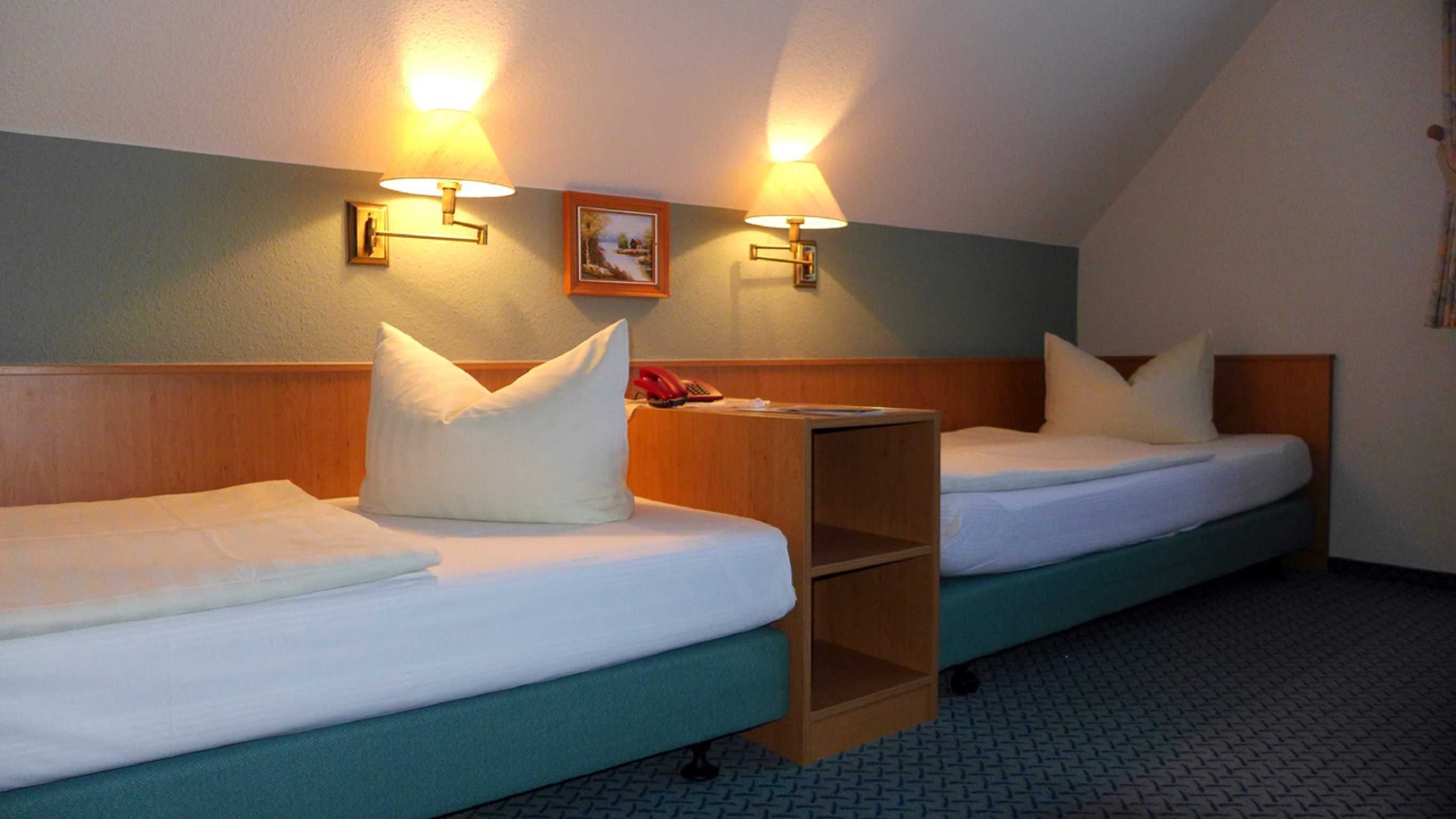 Habitación de hotel con dos camas individuales, cada una con cabecero de madera, ropa de cama blanca y una mesita de noche pequeña. Hay dos lámparas fijadas a la pared que proporcionan luz, y una pequeña estantería entre las camas.