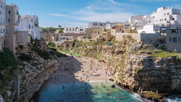 A coastal town with a rocky beach, clear blue waters, and white buildings.
