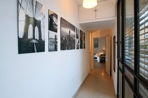 Un pasillo con una serie de fotografías blanco y negro en la pared, un accesorio de iluminación y una vista a través de una ventana con persianas.