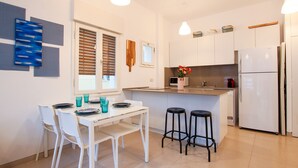 A modern kitchen with white cabinets, a central island with stools, a refrigerator, and a window with blinds. - MAMAD Trendy Apt & Balcony on Sheinkin Street by FeelHome (Tel Aviv - Jaffa)