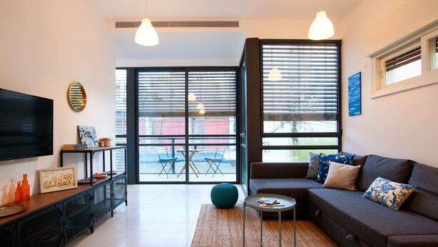 A modern living room with a sofa, a coffee table, and a TV. There is a balcony with a table and chairs outside. - MAMAD Trendy Apt & Balcony on Sheinkin Street by FeelHome (Tel Aviv - Jaffa)