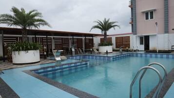 A swimming pool with lounge chairs and palm trees, surrounded by a building with a tiled roof and a balcony.