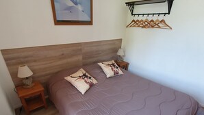 A bedroom with a wooden headboard, two bedside lamps, a bed with a purple quilt, and a wall-mounted coat rack. - Semi-Detached Chalet, 3 Rooms, 7 Pers, Terrace, Free Parking, Pets Allowed (La Joue du Loup)