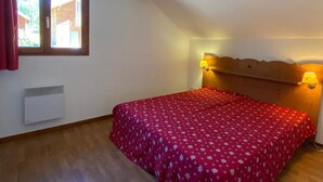 Une chambre avec un plafond en bois, un lit recouvert d’une courtepointe à motifs floraux rouges, deux lampes fixées au mur et une fenêtre avec des rideaux.