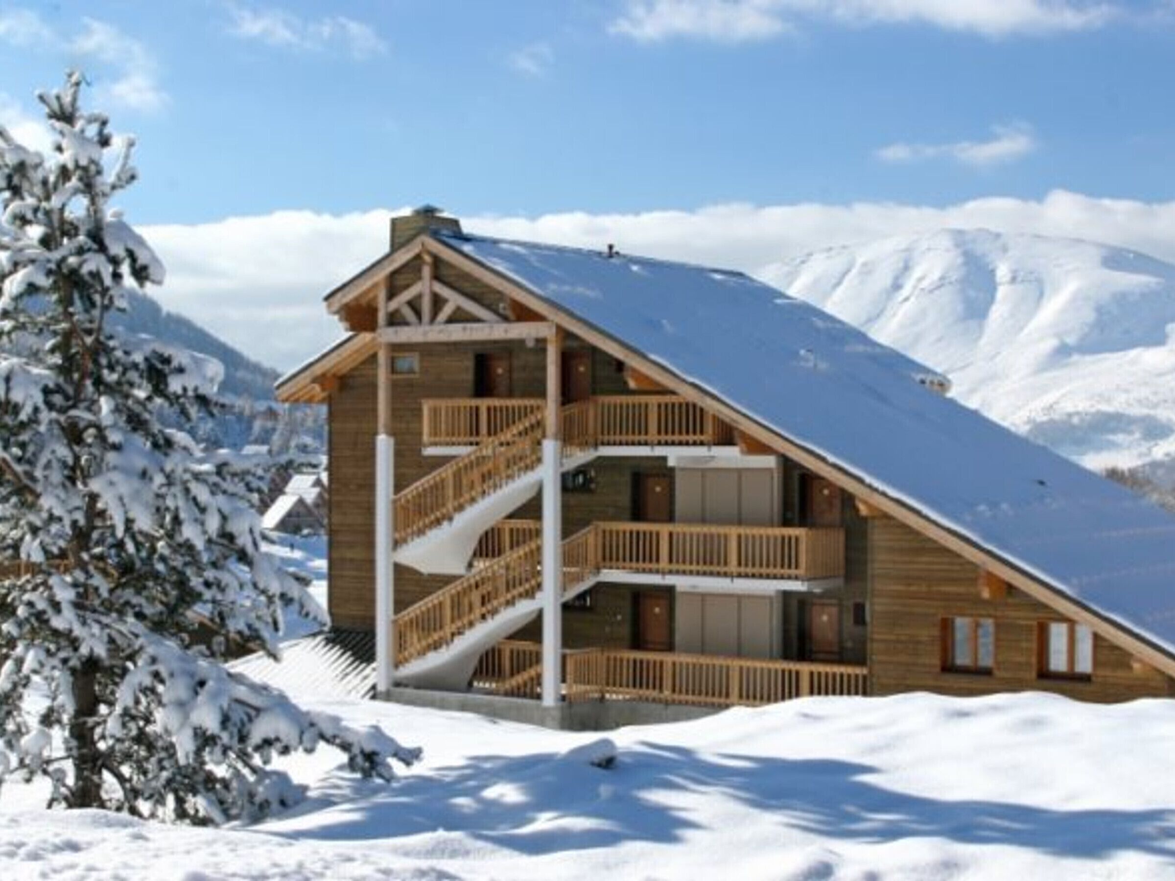 Un chalet en bois avec un toit pentu, un escalier et un balcon, entouré de montagnes enneigées.