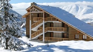 Un chalet en bois avec un toit pentu, un escalier et un balcon, entouré de montagnes enneigées.