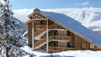 Un chalet en bois avec un toit pentu, un escalier et un balcon, entouré de montagnes enneigées.