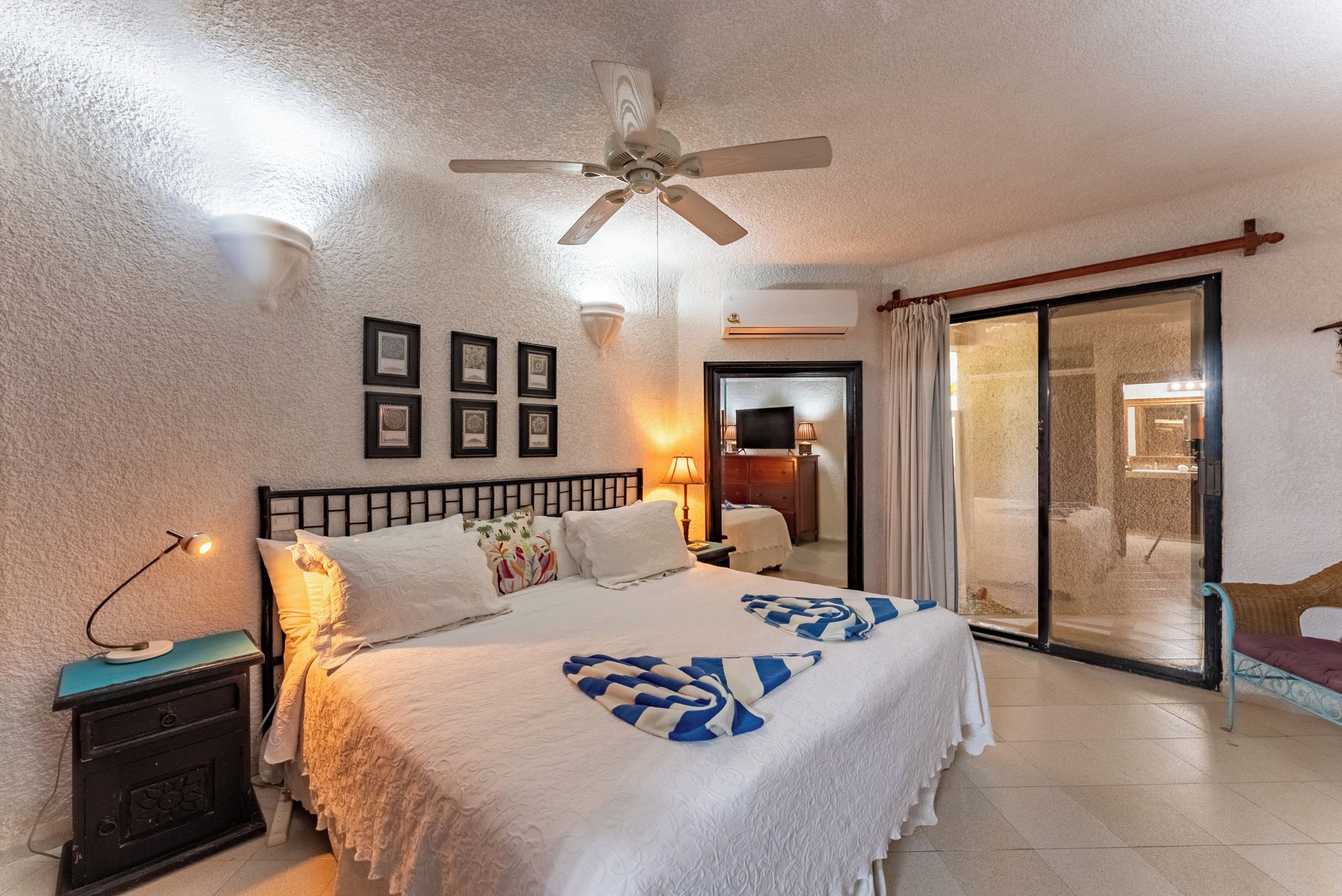 Quarto de hotel com cama, mesa de cabeceira, ventilador de teto, televisão e porta de vidro deslizante para uma varanda.
