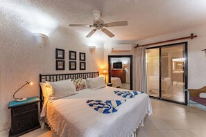 Habitación de hotel con cama, mesita de noche, ventilador de techo, televisor y puerta corrediza de vidrio que da a un balcón.