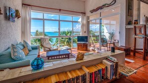 Ein geräumiges Wohnzimmer mit großem Fenster und Meerblick, eine Couch mit gemusterten Kissen, ein Couchtisch und ein Bücherregal voller Bücher.