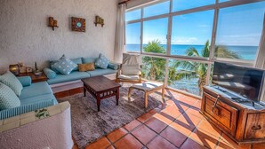 Ein Wohnzimmer mit großem Fenster und Meerblick, einer Couch, einem Sessel, einem Couchtisch und einem TV-Schrank.