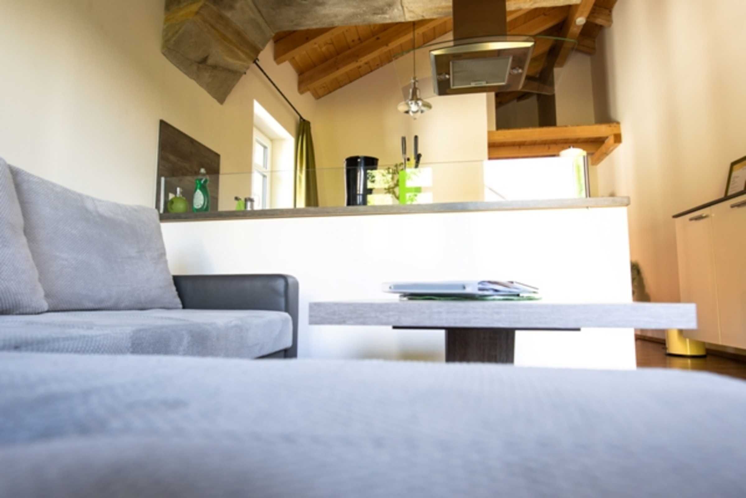 Ein modernes Wohnzimmer mit einem grauen Sofa, Holzboden und einer Küchenzeile im Hintergrund.